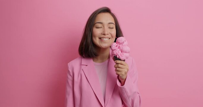 Pretty Asian woman gets pleasure from eating tasty appetizing ice cream licks delicious frozen dessert smiles happily enjoys spare time wears pink jacket poses indoor. Junk food much calories