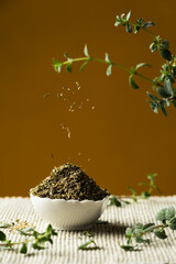 Heap of dried oregano in a white porcelain bowl, and shredded oregano falling, with fresh twigs and leaves, on a yellow or mustard color background, vertical image.