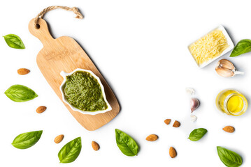 Basil pesto sauce in a white porcelain bowl on a small wooden table, with basil leaves, almonds, garlic, parmesan cheese and olive oil as ingredients on a white surface with copy space.
