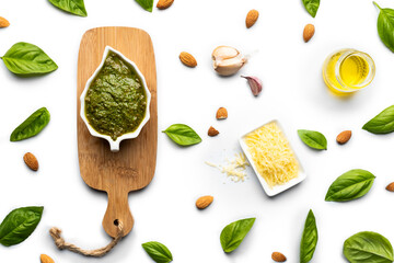 Delicious basil pesto sauce in a white porcelain bowl and a small wooden table, with basil leaves, almonds, garlic, parmesan cheese and olive oil as ingredients, on a white surface.