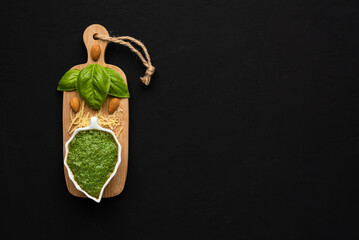 Basil pesto sauce in a leaf-shaped bowl on a small woden table with basil leaves, almonds and parmesan cheese on a black surface with space for text