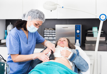 Professional woman cosmetician performing lifting procedure to mature woman on modern equipment in...