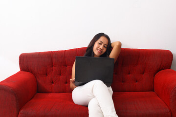 Obraz premium Asian woman using a laptop while sitting on red sofa.