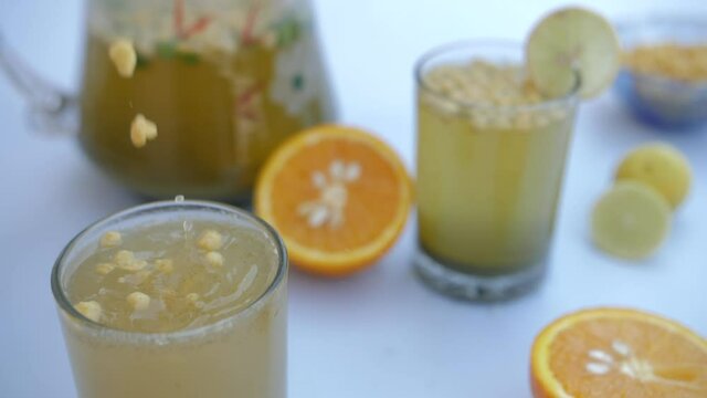 Crispy Boondi falling in a clear glass of refreshing Jaljeera drink - homemade recipe. Closeup shot of a jug half-filled with cool summer drinks  unpeeled oranges  and juicy lemon slices together