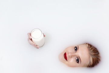 Attractive young woman with blue eyes and red lips lying in spa milk bath is holding glass of milk. Horizontally. 