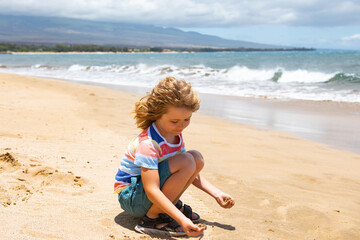 Cute boy playing with sand on summer tropical beach. Happy kid sit on the seaside sandy beach and playing with sand.