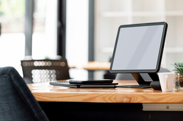 Mockup blank screen tablet on stand and gadget on wooden table in co-workspace.