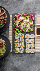 Mix of Asian dishes. Vegetable sushi maki rolls, gyoza dumplings with salad on a black stone board, pork and chicken dishes in bowls. Bamboo mat, nut and soy sauces. Top view. Copy space