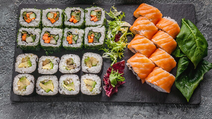 Food banner: set of different rolls on a black stone cutting board. Rolls with salmon, vegetables sushi maki rolls and fresh salad. Soy and peanut sauce, chopsticks. Gray concrete background. Top view