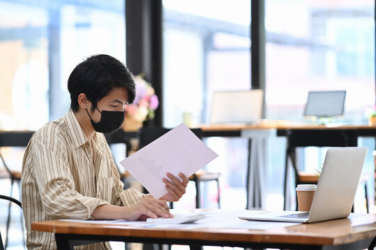 Young man office worker in protective mask analyzing business data at office desk. - Powered by Adobe