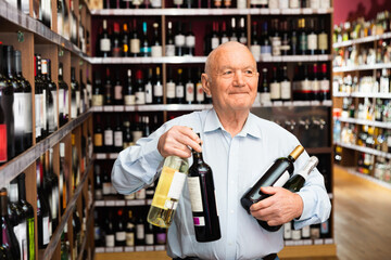 Cheerful positive mature man buying bottle of wine in store with alcohol drinks