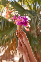 Woman holds bright pink flowers in an ice cream cone in front of of palm leaves in sunny weather. Advertising concept of travel and bright summer vacation. Summer surreal flowers creative and trendy. 