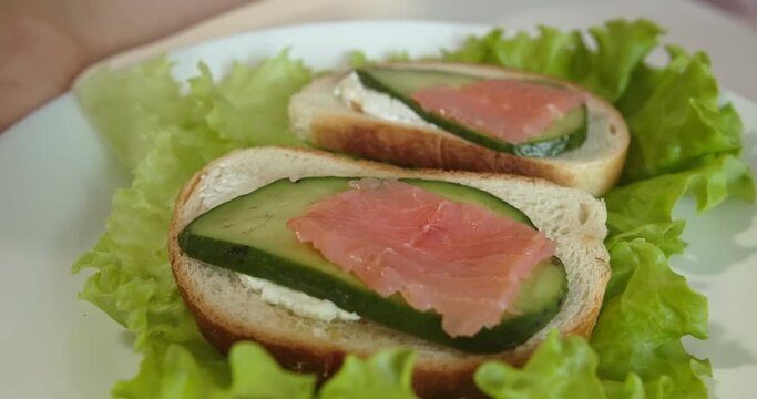 Two sandwiches on a plate close-up. They lie on lettuce leaves. This is white bread with butter and a slice of avocado. On one of them there is a salmon. The woman puts the same piece on the second
