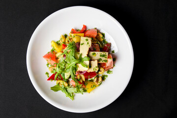 Vegetable salad with cheese, fresh romatoes and cucumbers, green olives served in a white plate over black background.