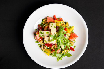 Vegetable salad with cheese, fresh romatoes and cucumbers, green olives served in a white plate over black background.