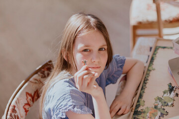 Cute little girl 8 -12 years old sitting at table holding one piece puzzle in hand and looking at camera. Brown-haired kid with long hair wearing casual blue dress.