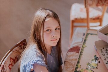Confident little girl 8 -12 years old sitting at table playing puzzle and looking at camera. Caucasian girl with long hair wearing casual blue dress. Leisure activity at home.