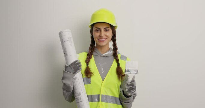 Pleased female architect holds painting brush for refurbishment paper blueprint wears protective hardhat uniform and gloves isolated over white background. Home repair redecorating and construction