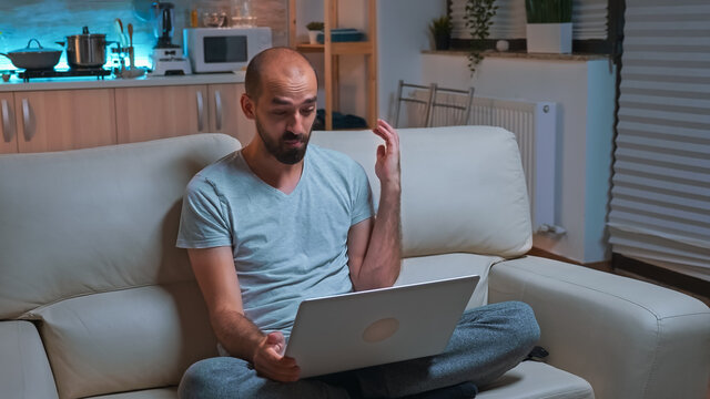 Focused Man Working On Laptop Computer During Business Videocall Conference. Caucaisan Male Sitting On Sofa In Pajamas Late At Night In Kitchen Using Modern Technology Wireless