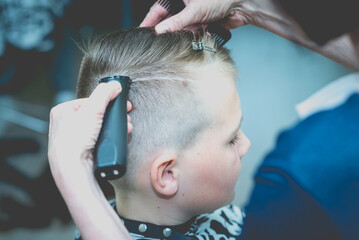 Hairdresser makes a fashionable pretty hairstyle for the young boy in a modern barbershop.Side...