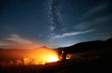 Fantastic view of night starry sky over grassy hill with male traveler near campfire. Man hiker sitting on wooden plank near bright bonfire and looking at beautiful sky with stars and Milky Way.