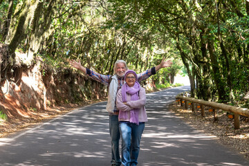 Smiling senior couple with raised arms looking at camera enjoying mountain excursion in a cool day. Active retired elderly people and fun concept