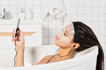 relaxed young woman with sheet mask on face lying in bathtub with earphones and cellphone in hand