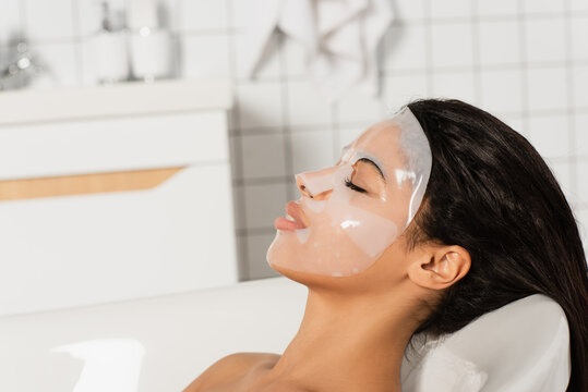 young african american woman with sheet mask on face lying in bathtub