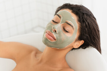 relaxed young african american woman with mud mask on face lying in bathtub