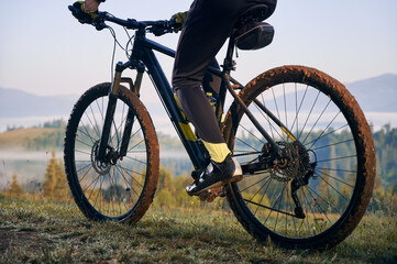 Fototapeta na wymiar Horizontal cropped snapshot of biker's legs, sitting on bicycle. Close-up view on bike, wheels in mud, after morning ride on mountains trails. Athlete enjoying the moment of sunrise
