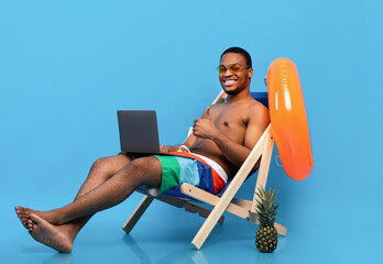 Cheerful black guy using laptop, browsing internet during beach vacation, sitting in lounge chair,...