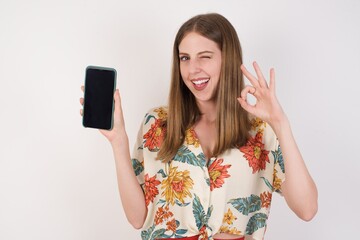 Excited woman showing smartohone blank screen, blinking eye and doing ok sign with hand. Studio shot of shocked girl holding smartphone with blank screen. Advertisement concept.