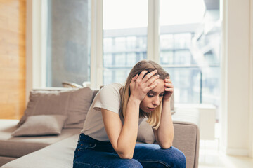 Portrait young woman sitting on sofa at home depressed. Girl is having a hard time with stress. Caucasian female indoors alone worried about problems. unhappy blonde student in living room or bedroom
