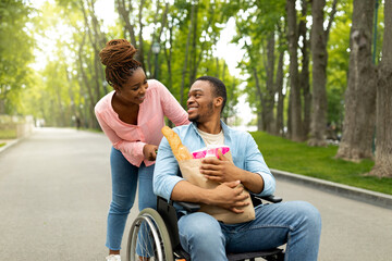 Happy young impaired man in wheelchair carrying paper bag with food, his lovely wife helping him...