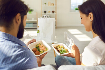Happy people enjoying takeout lunch at home. Young couple sitting on sofa and eating fresh healthy takeaway food from plastic containers. Man and woman comparing two meals ordered in delivery service