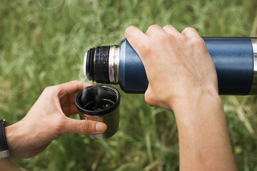 Male hands holding a thermos and pouring hot tea outdoors, close-up. Hike, camping on the nature...