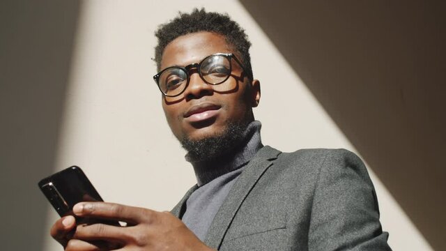 Low angle portrait shot of young handsome Afro-American businessman in stylish formalwear and glasses typing on smartphone and then looking at camera and smiling