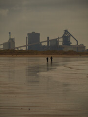 Bleached, stark landscape, a dull sky reflected on wet sand, two people silhouetted in the mid ground and a near-silhouetted steel mill on the horizon.