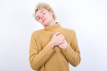 Honest Young handsome Caucasian blond man standing against white background keeps hands on chest,...