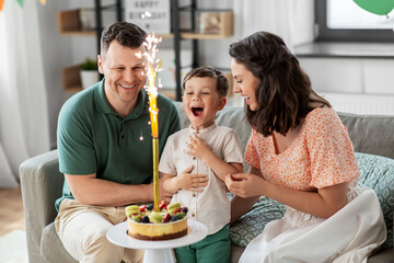 family, holidays and people concept - portrait of happy mother, father and little son with firework candle burning on birthday cake sitting on sofa at home party