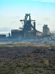 The derelict, abandoned hulk of the blast furnace at the Redcar Steelworks in a haze against a light blue sky, seen from the rubble of the slagfields.