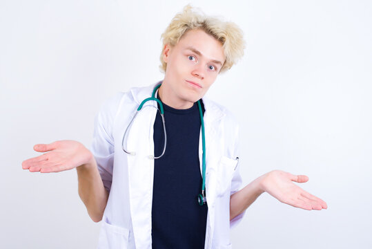 Puzzled And Clueless Young Handsome Caucasian Doctor Man Standing Against White Background With Arms Out, Shrugging Shoulders, Saying: Who Cares, So What, I Don't Know. Negative Human Emotions.