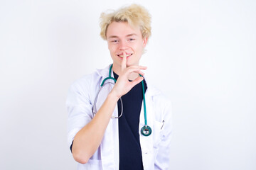 Smiling young handsome Caucasian doctor man standing against white background makes shush gesture,...