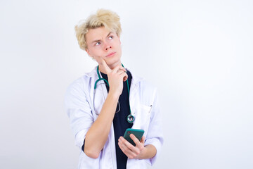 Thinking dreaming young handsome Caucasian doctor man standing against white background using mobile phone and holding hand on face. Taking decisions and social media concept.