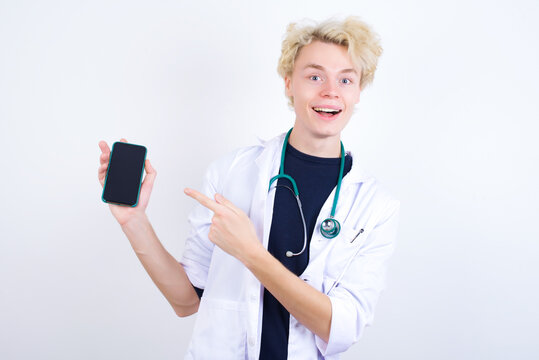 Excited young handsome Caucasian doctor man standing against white background holding and pointing with finger at smartphone with blank screen. Advertisement concept.