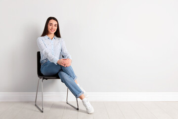 Young woman sitting on chair near white wall in office, space for text