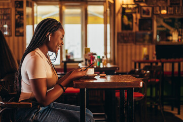 Cheerful african american woman using a smartphone and airpods while sitting in a cafe