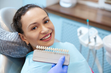 Brunette woman with beautiful smile before receiving dental care check up and teeth whitening bleaching, dentist wearing exam gloves checks tooth color with a comparison veneer scale chart.