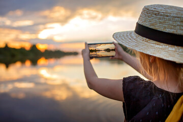 Pretty preteen girl in summer straw hat taking selfie for cellphone at beautiful sunset on lake,...