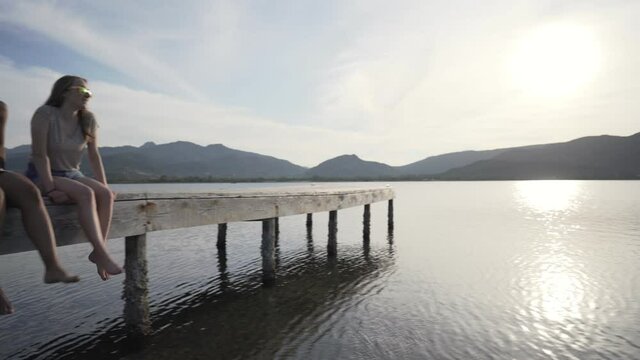 Multiracial happy gen z friends group sitting with dangling feet on a pier at sunset or dawn looking each other. Camera pan on sea ocean or flat lake water with sun reflection effect. People on jetty